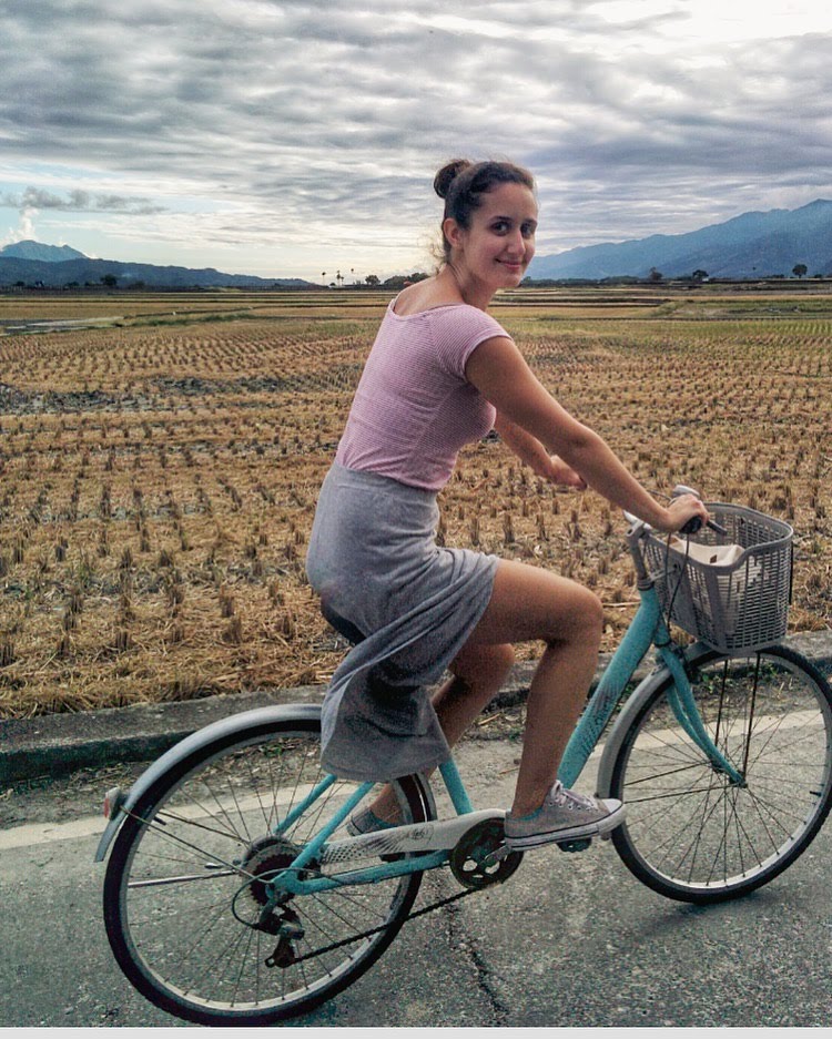 Vivian Trumblay riding on a bike in a rural area, next to a field with mountains in the distance.