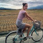 Vivian Trumblay riding on a bike in a rural area, next to a field with mountains in the distance.